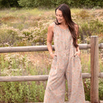 Woman wearing a floral printed gray jumpsuit with boots, standing in front of a fence and trees and bushes