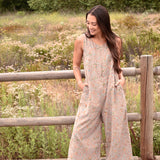 Woman wearing a floral printed gray jumpsuit with boots, standing in front of a fence and trees and bushes