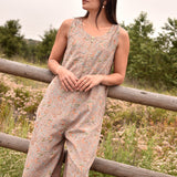 Angled shot of woman in block printed floral printed gray sleeveless jumpsuit, woman is leaning against a fence