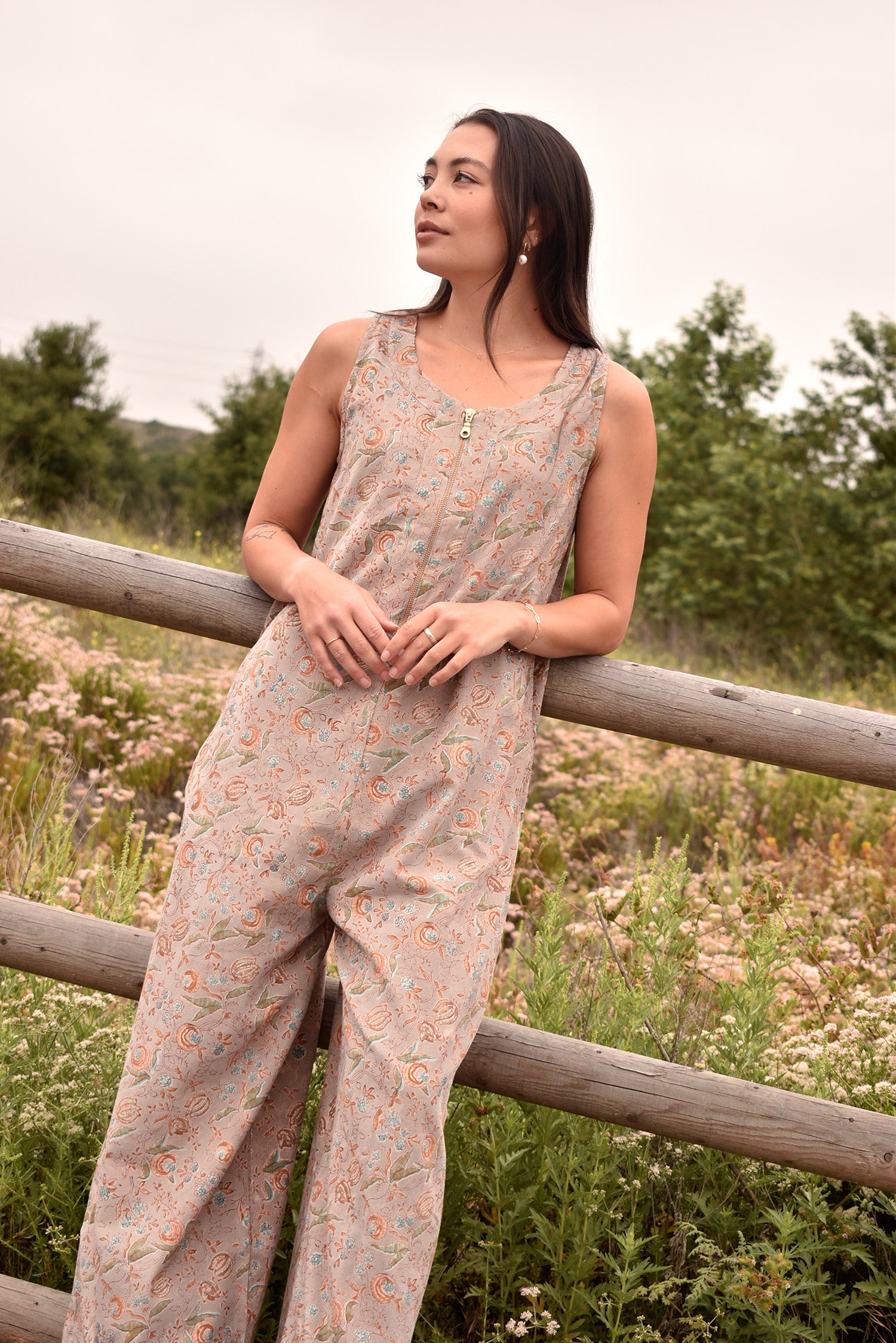 Angled shot of woman in block printed floral printed gray sleeveless jumpsuit, woman is leaning against a fence