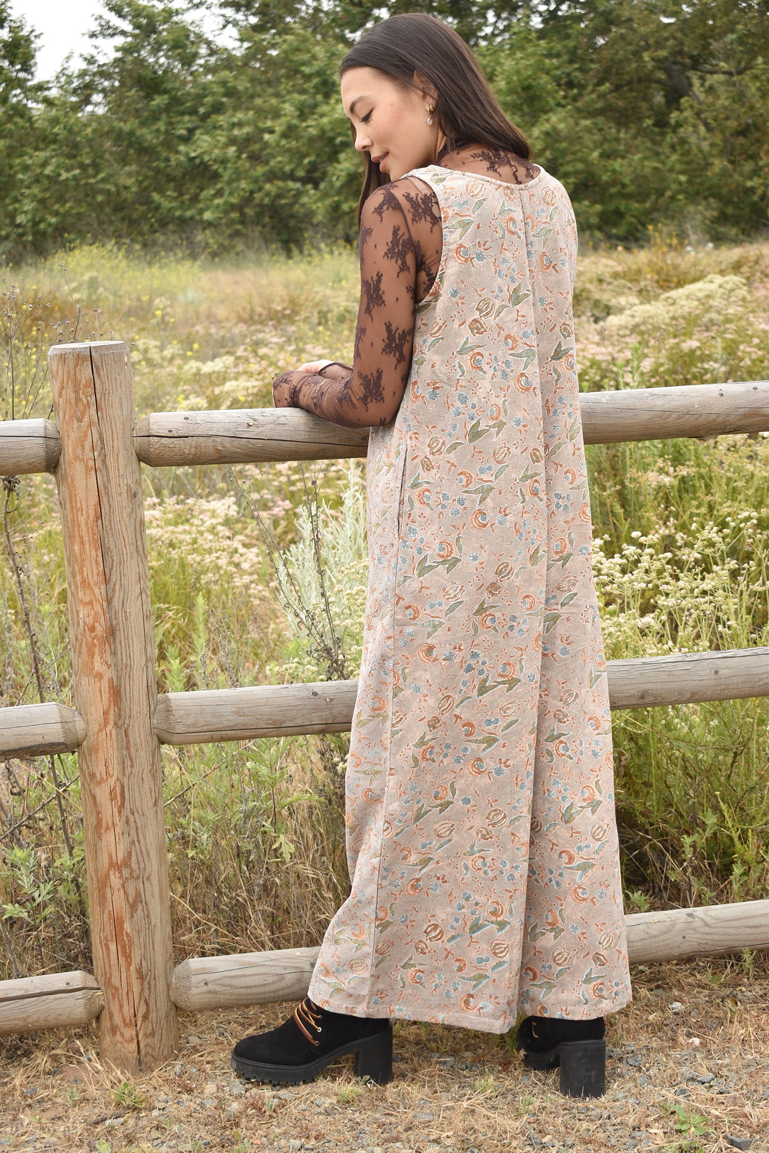 Back shot of woman wearing a sheer lace long sleeve under block printed floral printed gray jumpsuit. Woman it outside standing in front of a fence in nature.