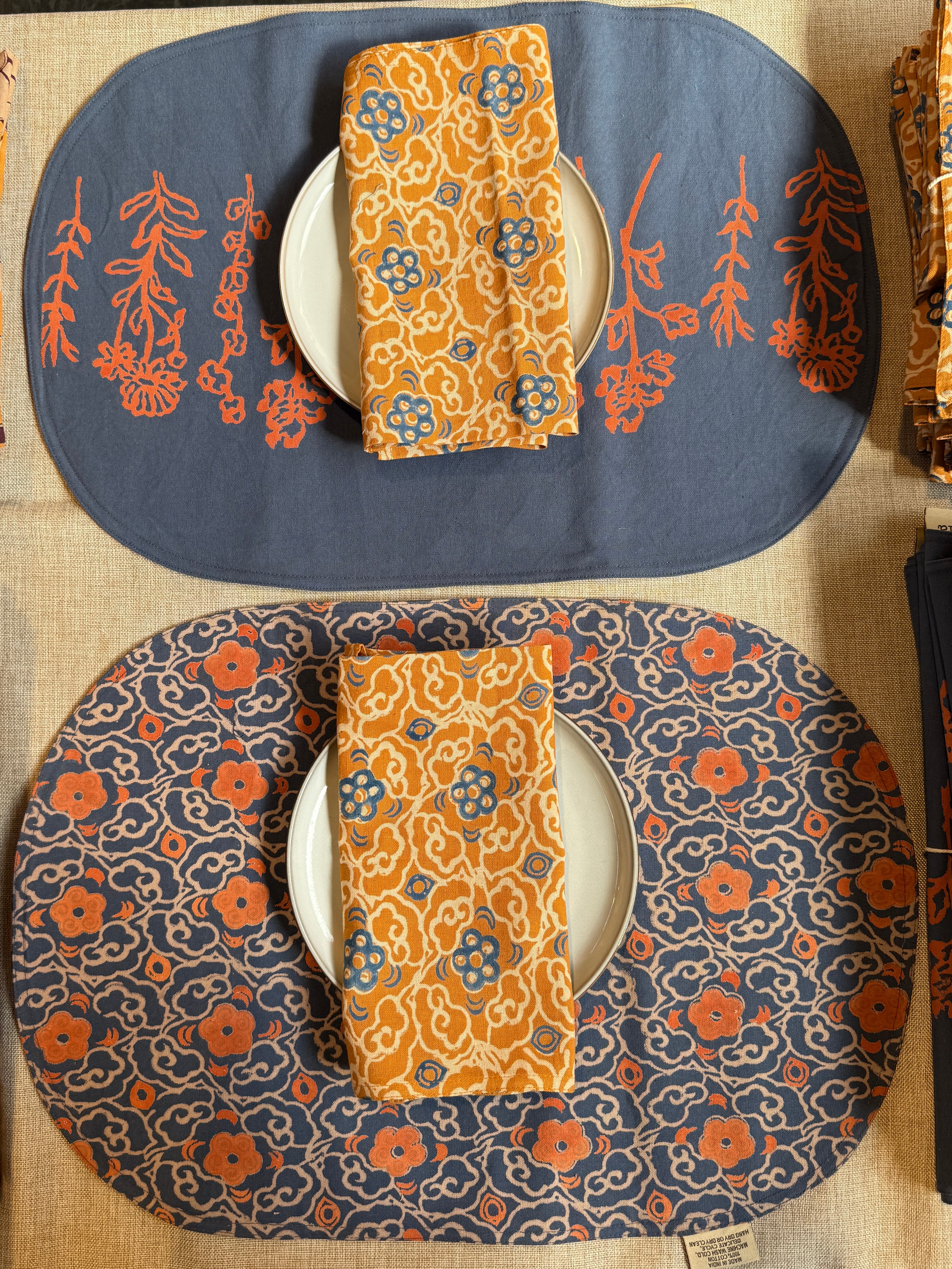 Two Marigold placemats set on a table, one showing the front print and the other showing the back print. Each styled with a plate and folded Marigold napkin on top of the plate
