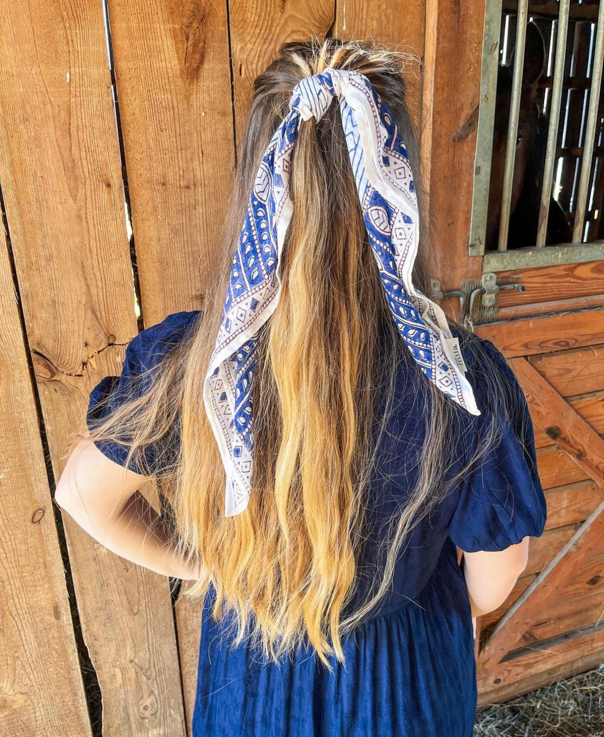 Woman wearing a blue printed scarf styled in hair. Woman is wearing a blue dress standing in front of a barn.