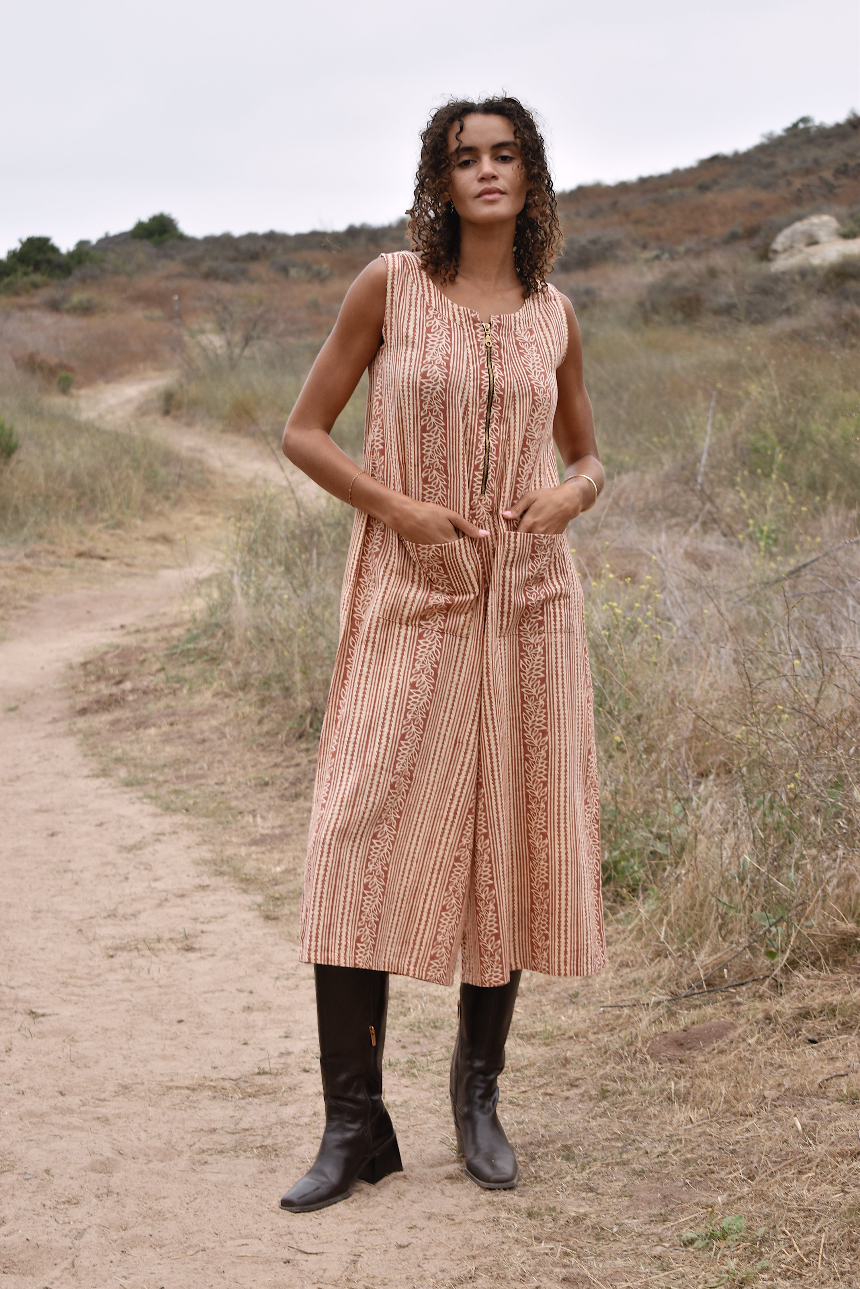 Model wearing sleeveless, cropped jumpsuit outside with heeled brown boots. Jumpsuit has rust floral print, two front pockets and front zipper.