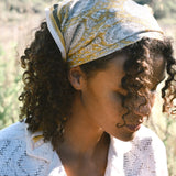 Woman wearing block print Tate bandana wrapped around the hair. Woman wearing a white, collared shirt. Foliage in the background.