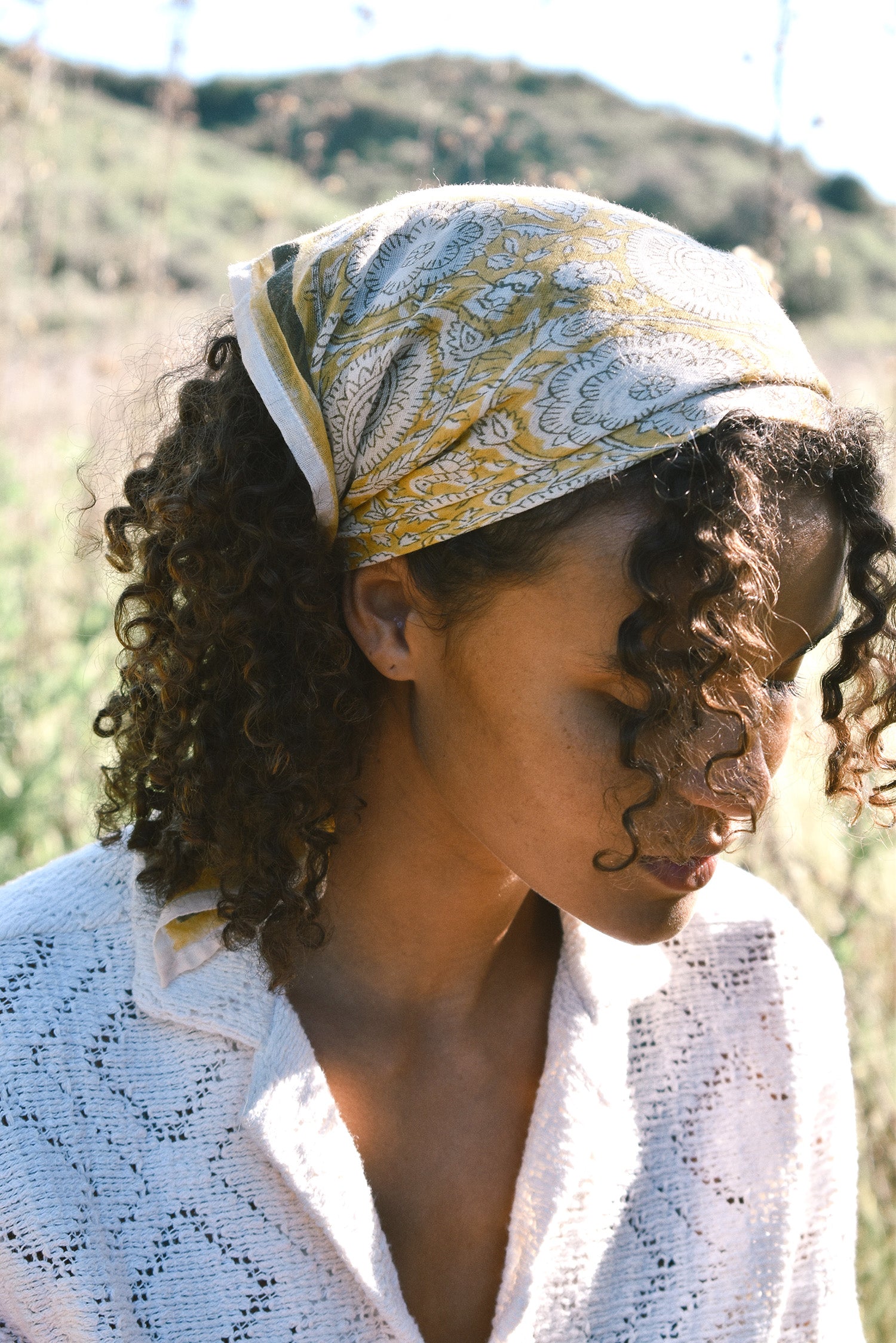 Woman wearing block print Tate bandana wrapped around the hair. Woman wearing a white, collared shirt. Foliage in the background.