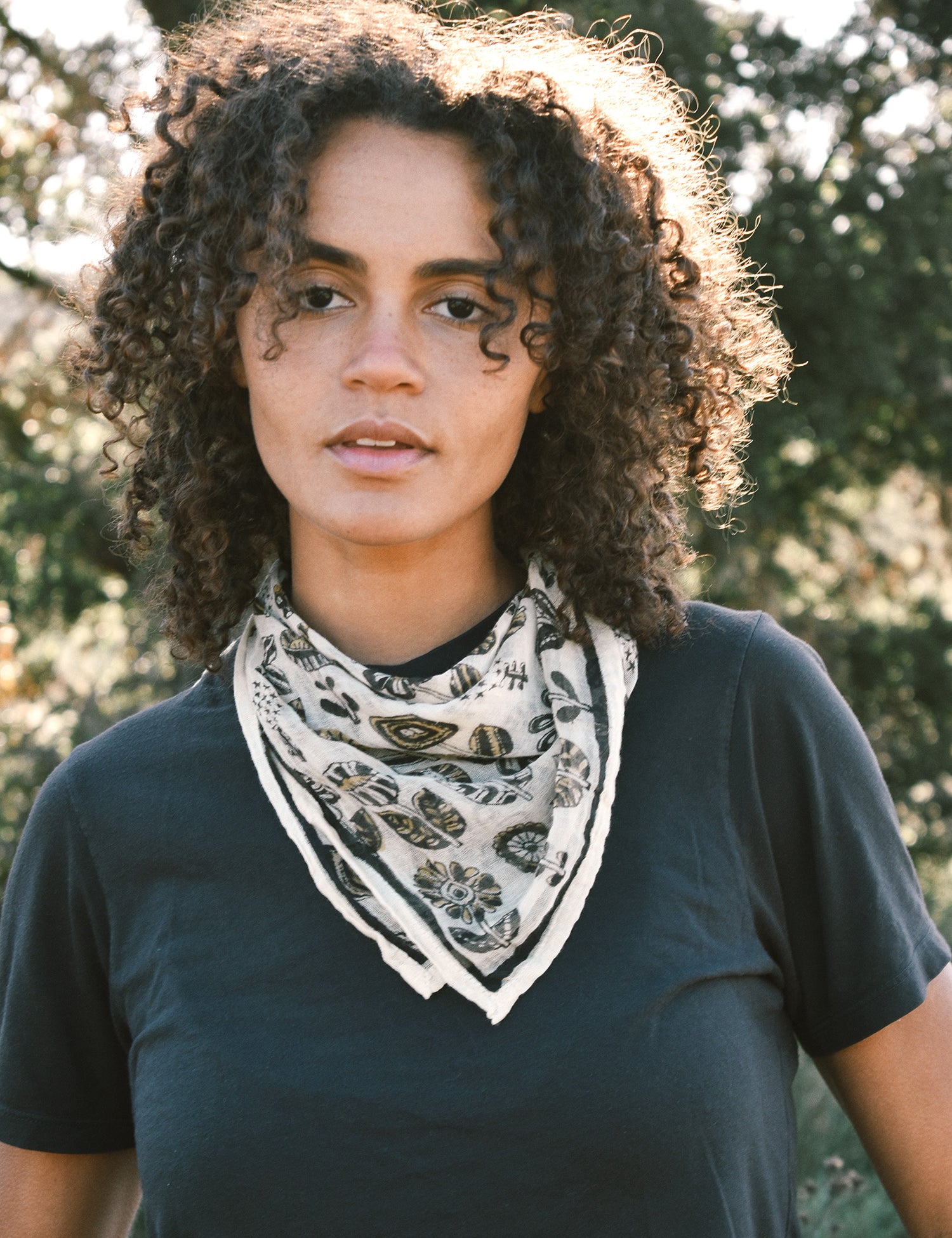 Woman wearing block print Paradise bandana tied around neck against a black tee shirt. Trees are in the background.