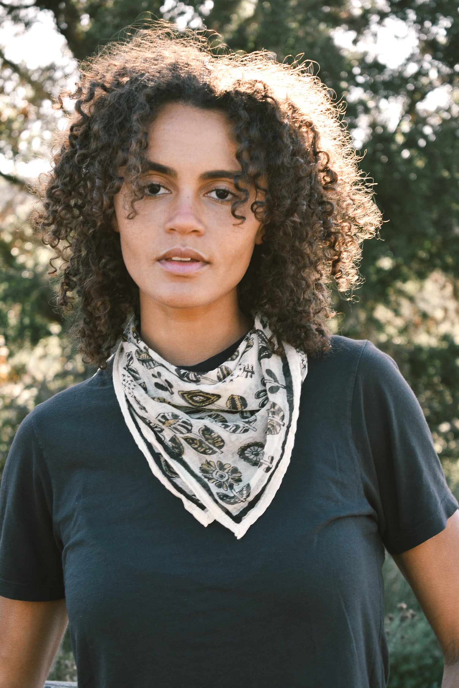 Woman wearing block print Paradise bandana tied around neck against a black tee shirt. Trees are in the background.