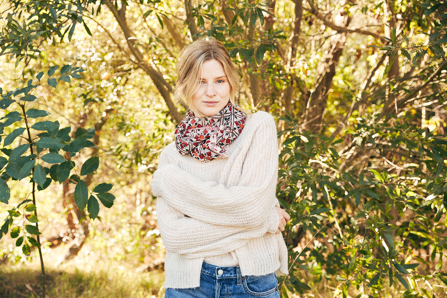 Woman wearing a scarf tied around the neck with a white sweater, blue jeans. Trees in the background.