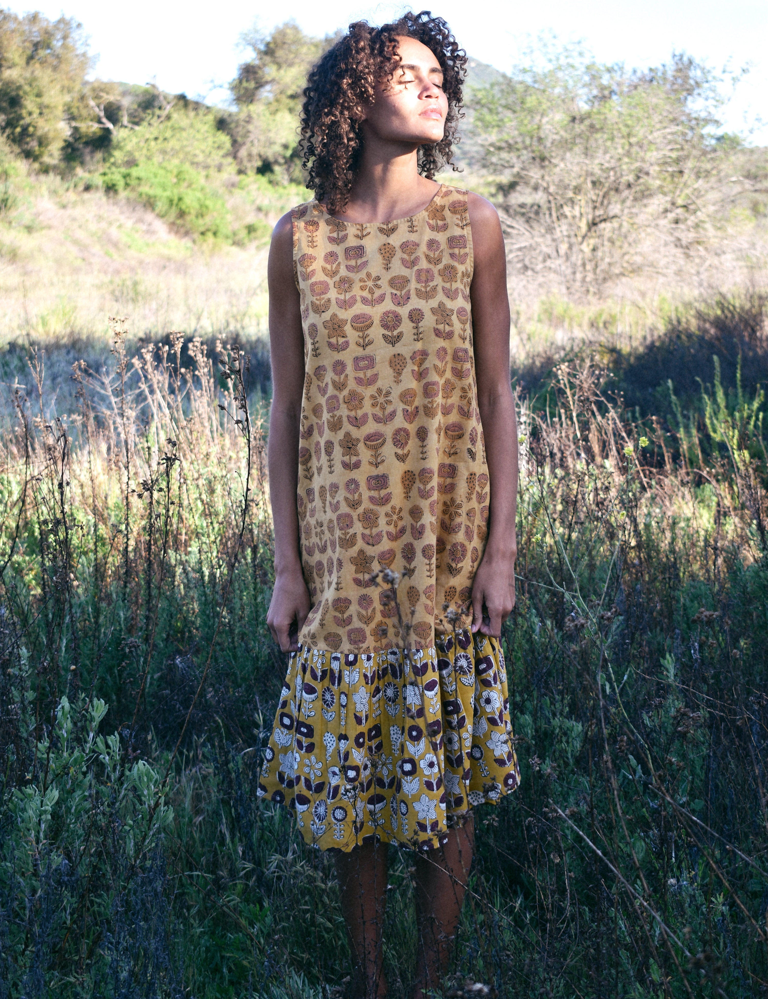 Woman wearing Sharon dress, standing in a field of tall grass. Woman looking up towards the sun.