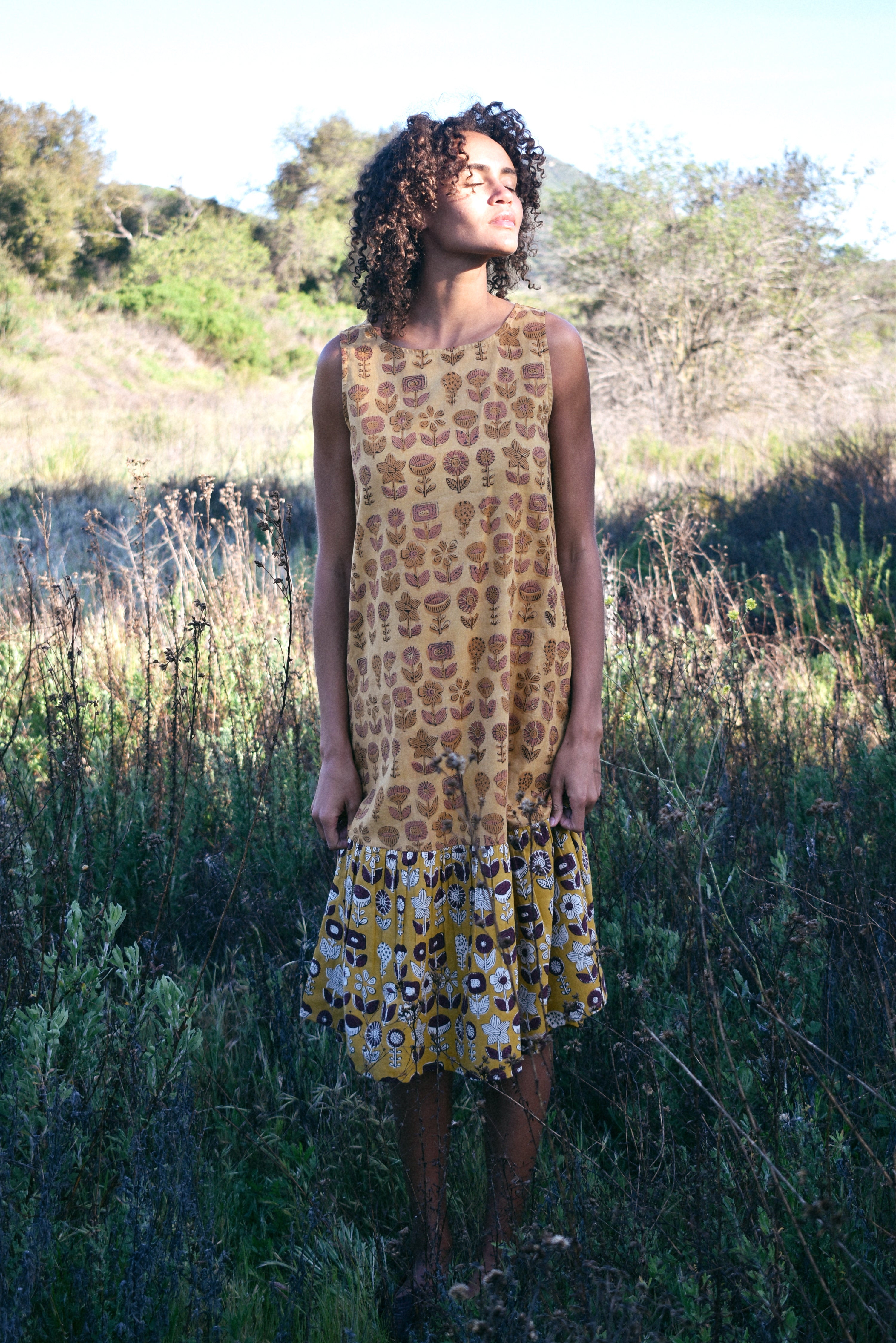 Woman wearing Sharon dress, standing in a field of tall grass. Woman looking up towards the sun.