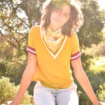 Woman wearing block print Tate bandana around the neck against a yellow tee shirt. Woman sitting on wooden fence with trees in the background.