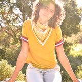 Woman wearing block print Tate bandana around the neck against a yellow tee shirt. Woman sitting on wooden fence with trees in the background.