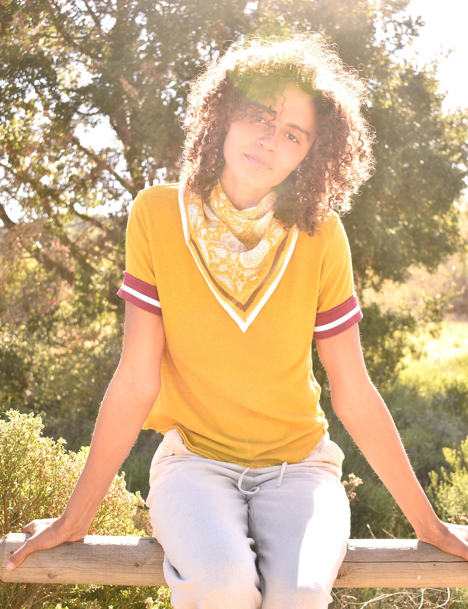Woman wearing block print Tate bandana around the neck against a yellow tee shirt. Woman sitting on wooden fence with trees in the background.