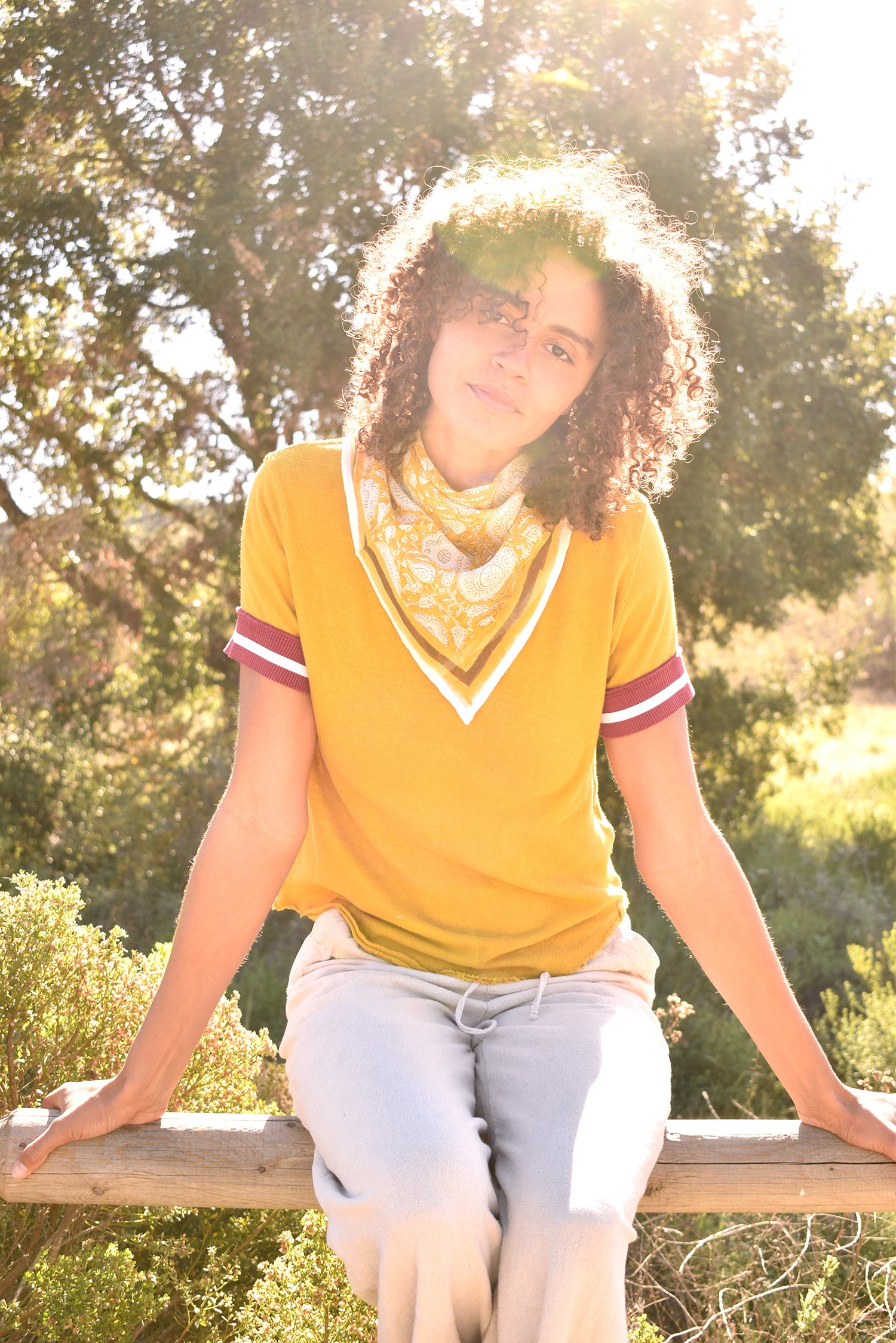 Woman wearing block print Tate bandana around the neck against a yellow tee shirt. Woman sitting on wooden fence with trees in the background.