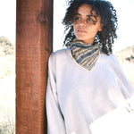 Woman wearing block print Frontier bandana tied around neck against a grey sweatshirt. Woman leaning against wooden pillar.