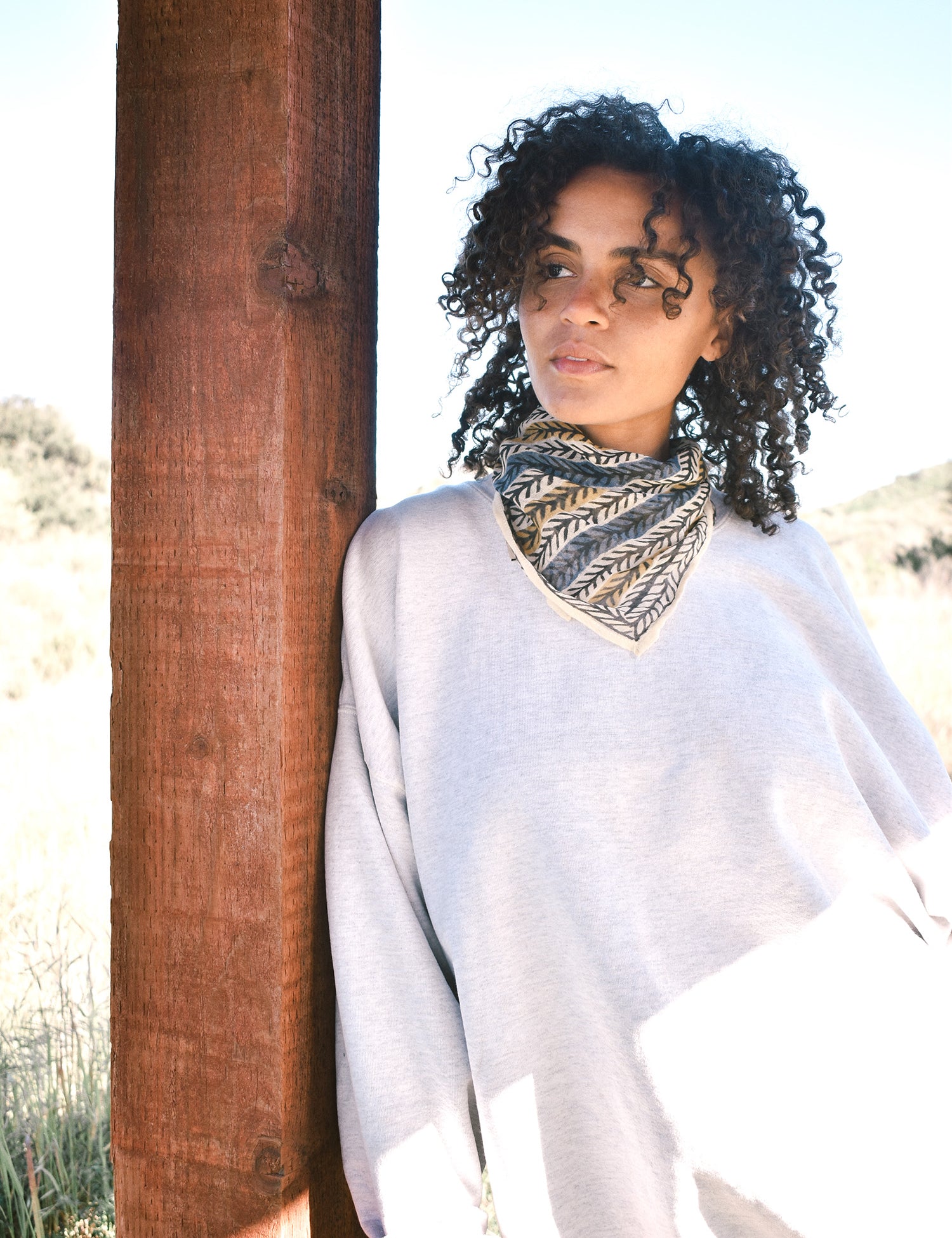 Woman wearing block print Frontier bandana tied around neck against a grey sweatshirt. Woman leaning against wooden pillar.