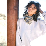 Woman wearing block print Frontier bandana tied around neck against a grey sweatshirt. Woman leaning against wooden pillar.