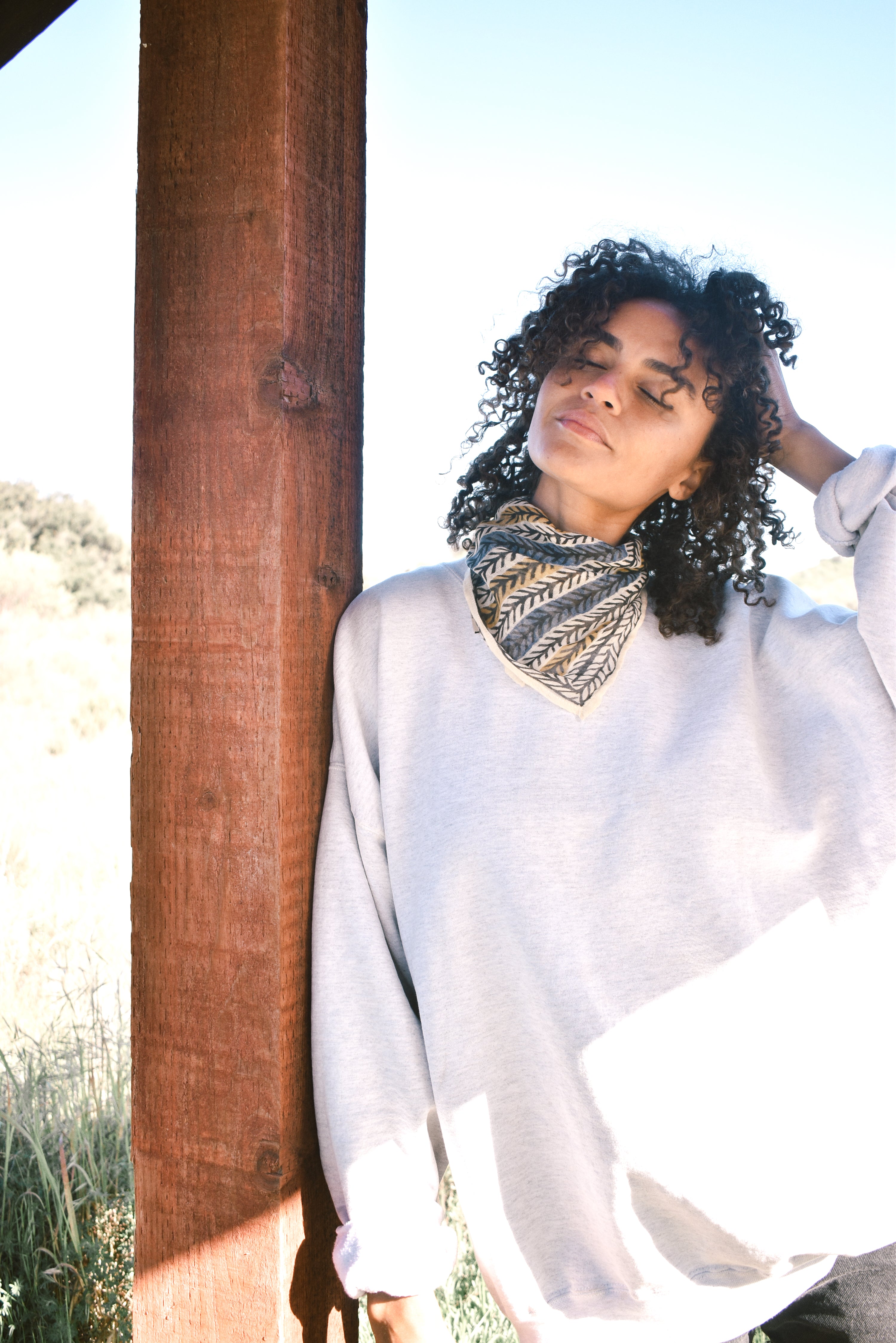 Woman wearing block print Frontier bandana tied around neck against a grey sweatshirt. Woman leaning against wooden pillar.