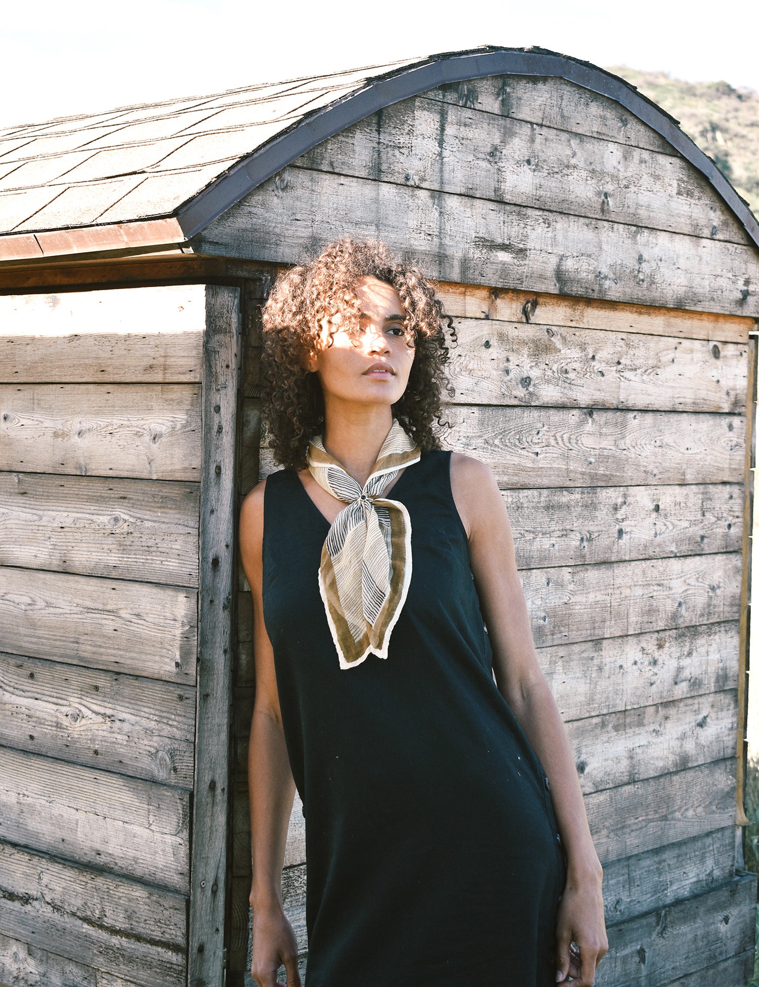 Woman wearing block print Savanna bandana tied around neck against a black sleeveless dress. Woman leaning against a wooden building.