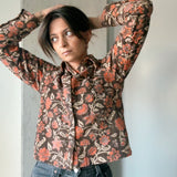 Woman wearing block printed floral printed corduroy shirt with black jeans, standing in front of wall. Woman's hand is in in her hair.