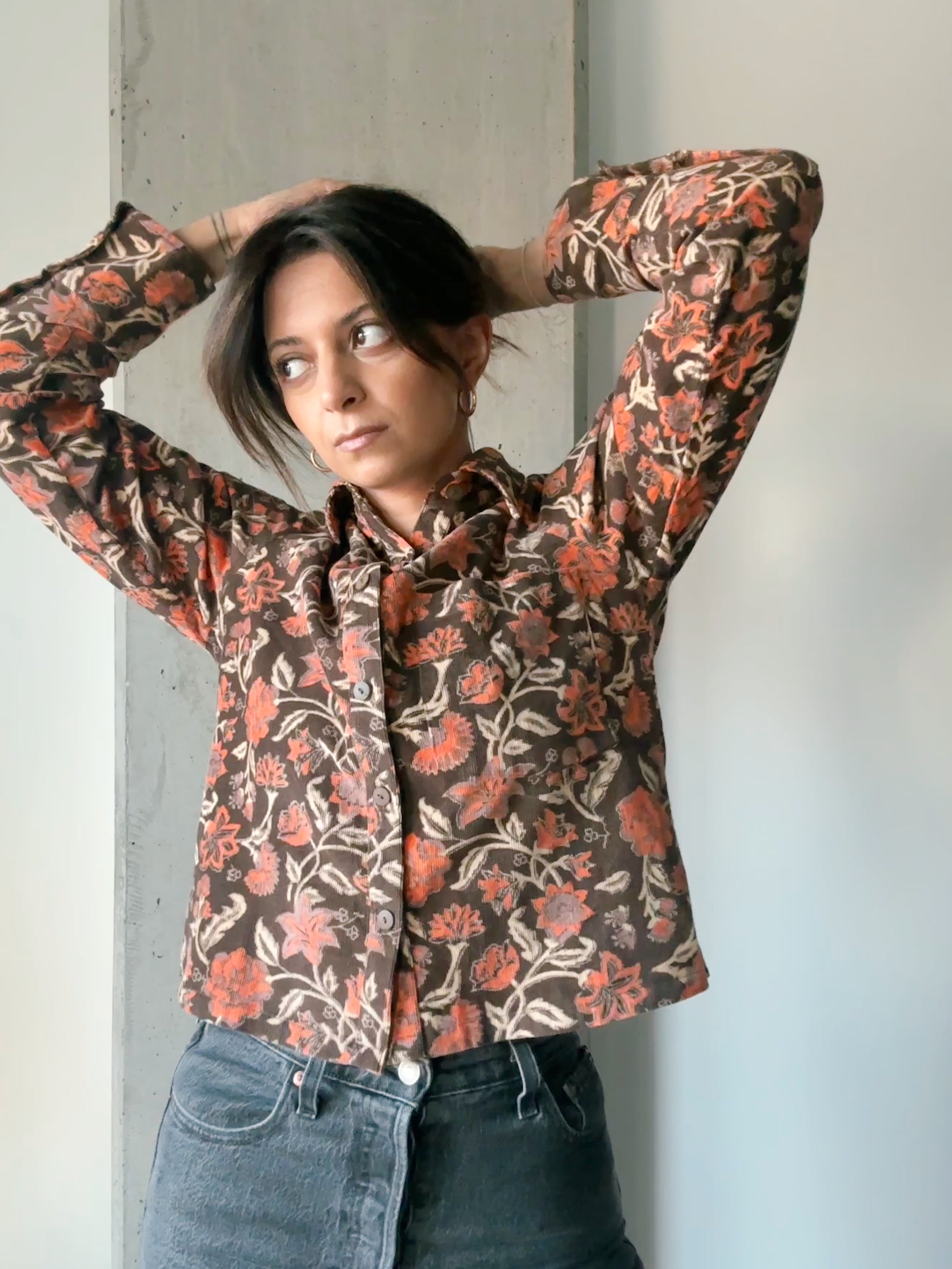 Woman wearing block printed floral printed corduroy shirt with black jeans, standing in front of wall. Woman's hand is in in her hair.