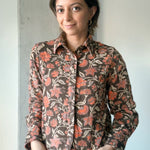 Woman is wearing block printed, brown floral printed corduroy shirt with jeans. Woman is standing in front of a white and grey wall.