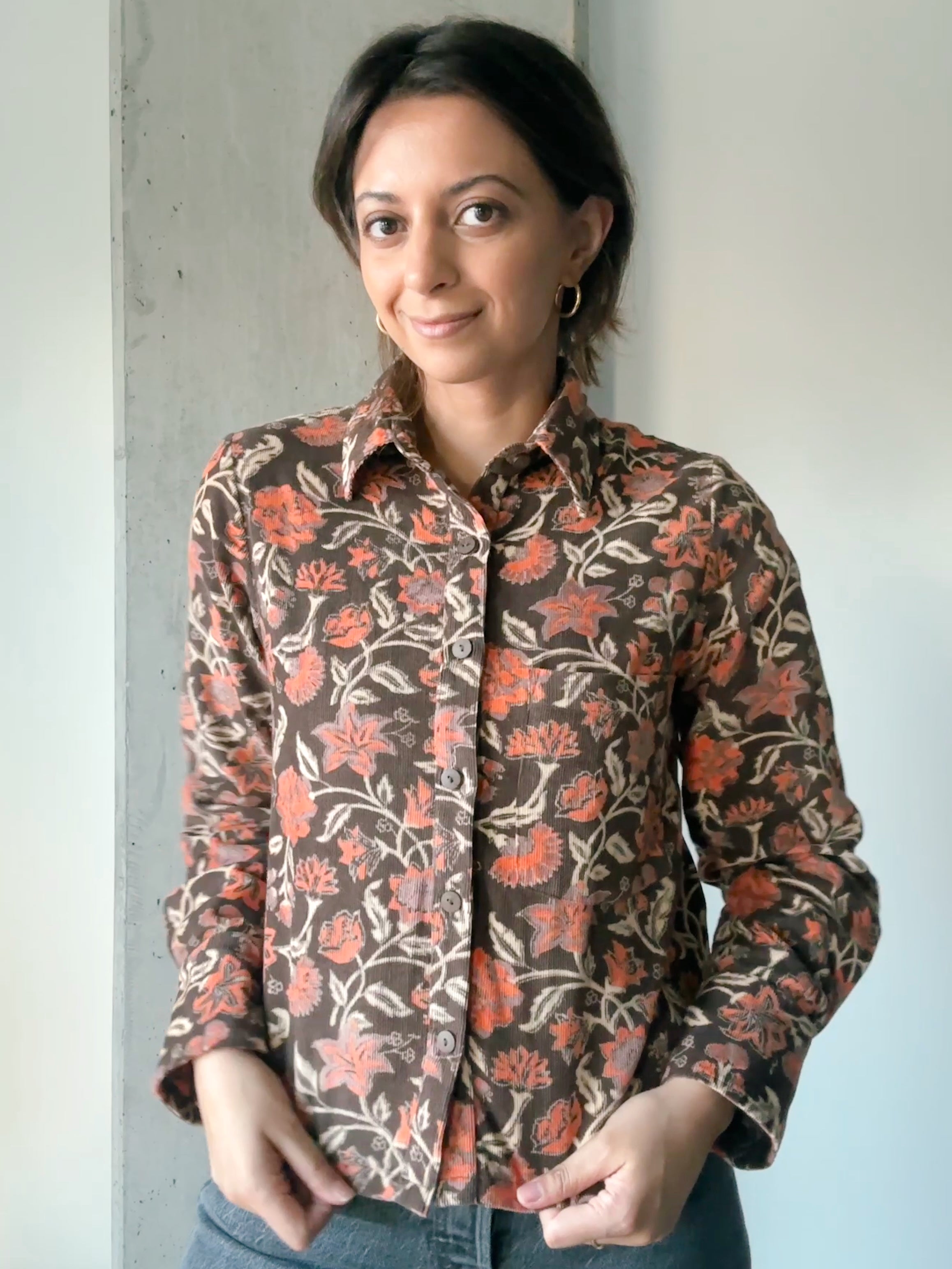 Woman is wearing block printed, brown floral printed corduroy shirt with jeans. Woman is standing in front of a white and grey wall.
