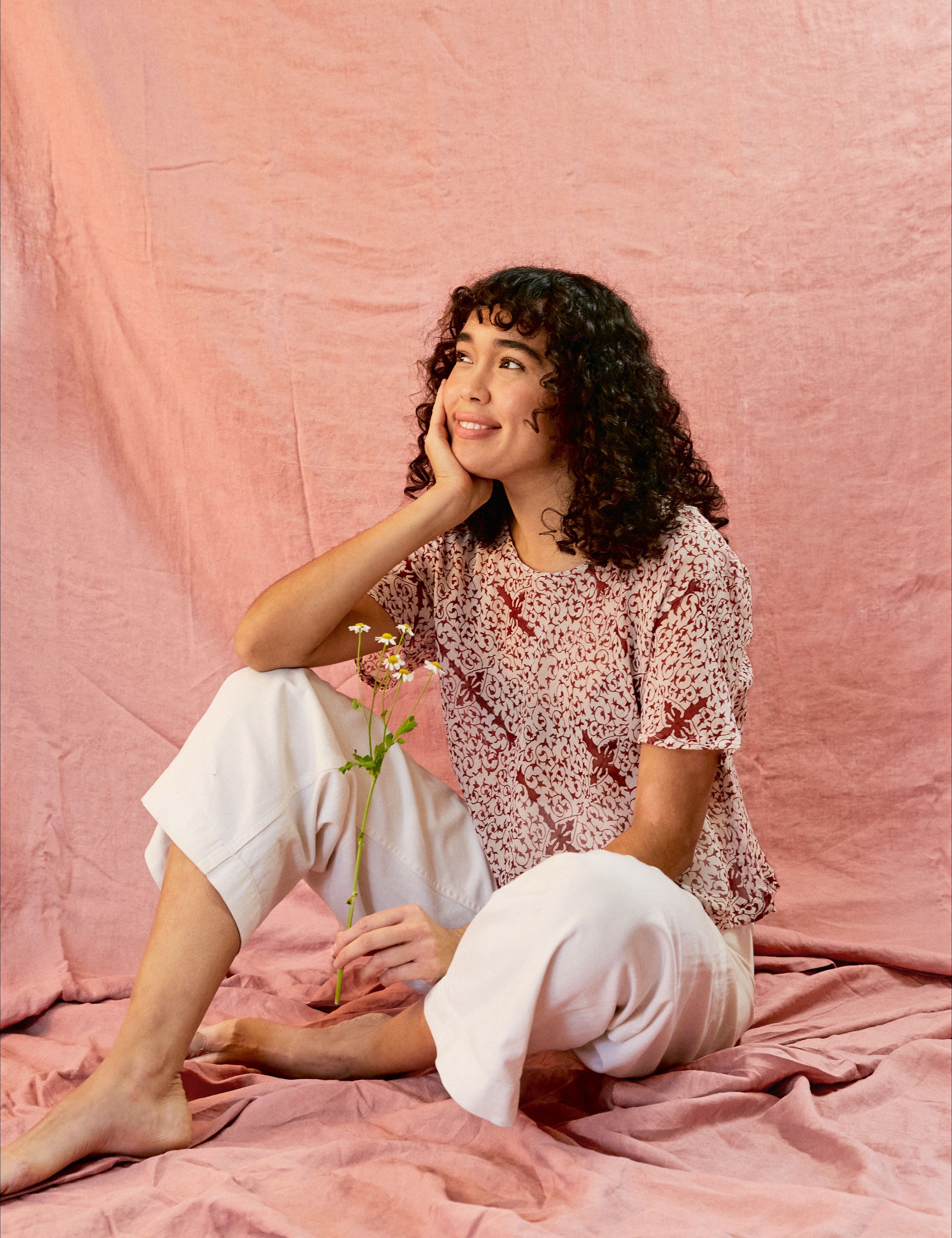 Model wearing the Alicia Top with white pants, against a pink backdrop. Model is holding a flower and sitting down.