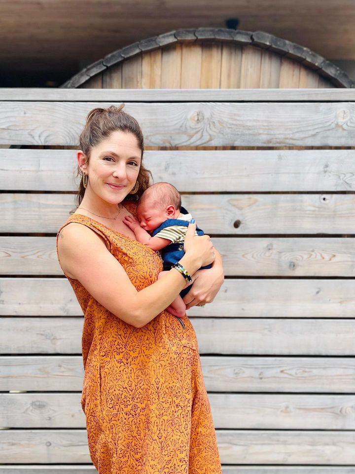 New mom holding a baby while wearing a block printed orange jumpsuit with front zipper opening