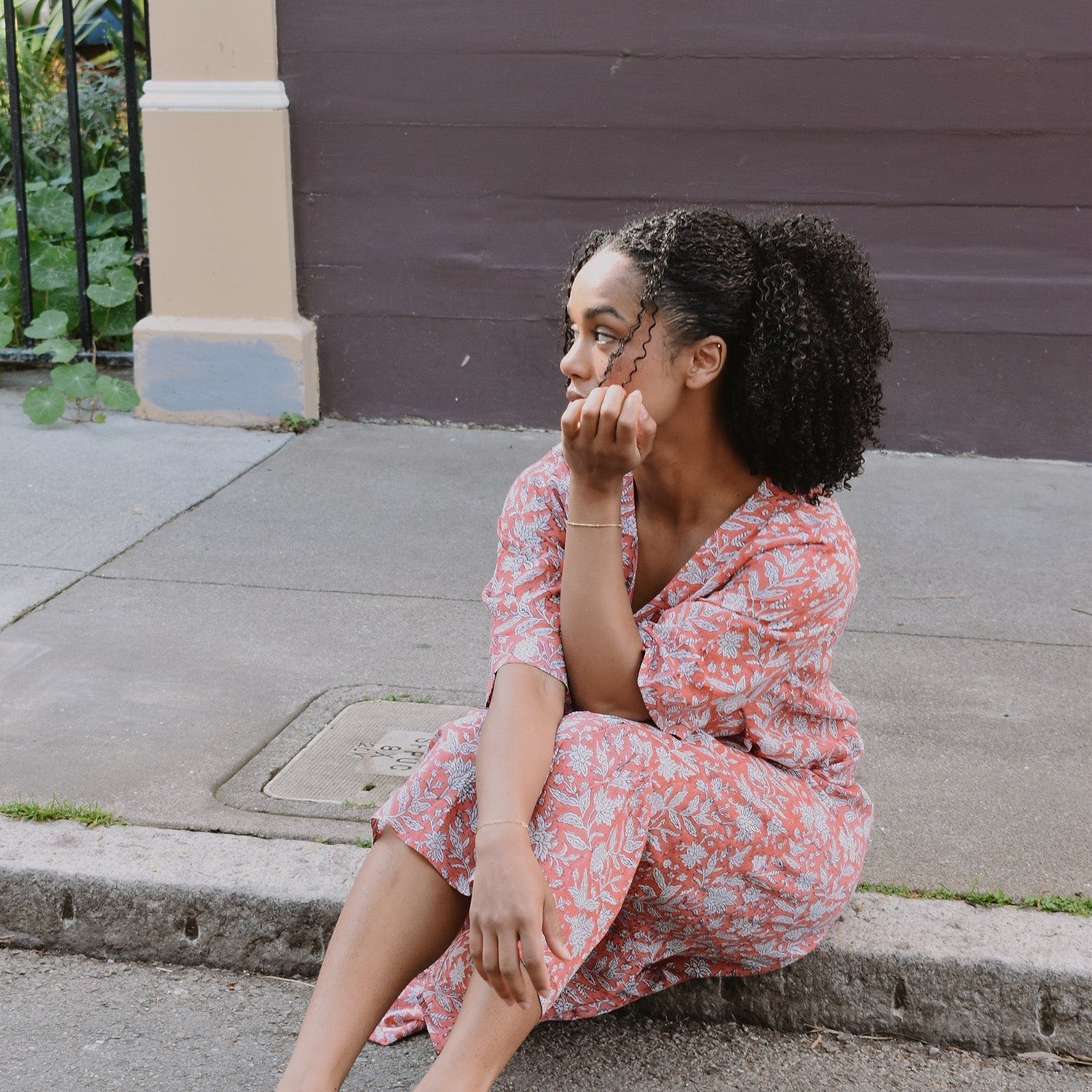 Block printed pink floral summer midi ruffle dress. Woman wearing dress and sitting on a curb
