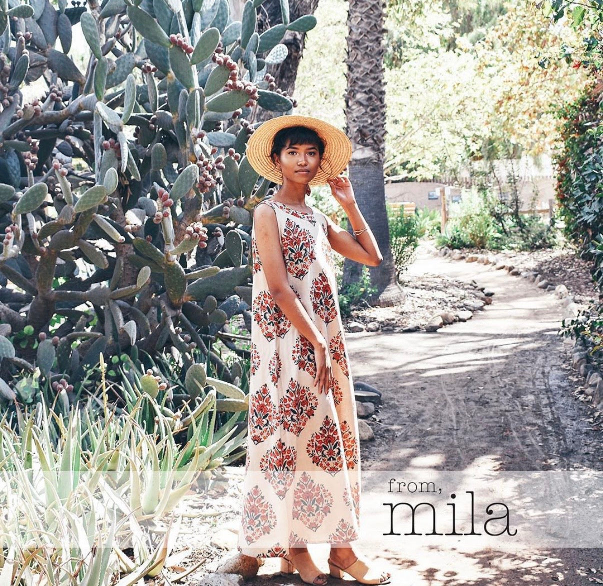 Woman in a sleeveless block printed maxi dress and straw sun hat standing on a path beside cactus and desert plants, From Mila logo overlay in corner.