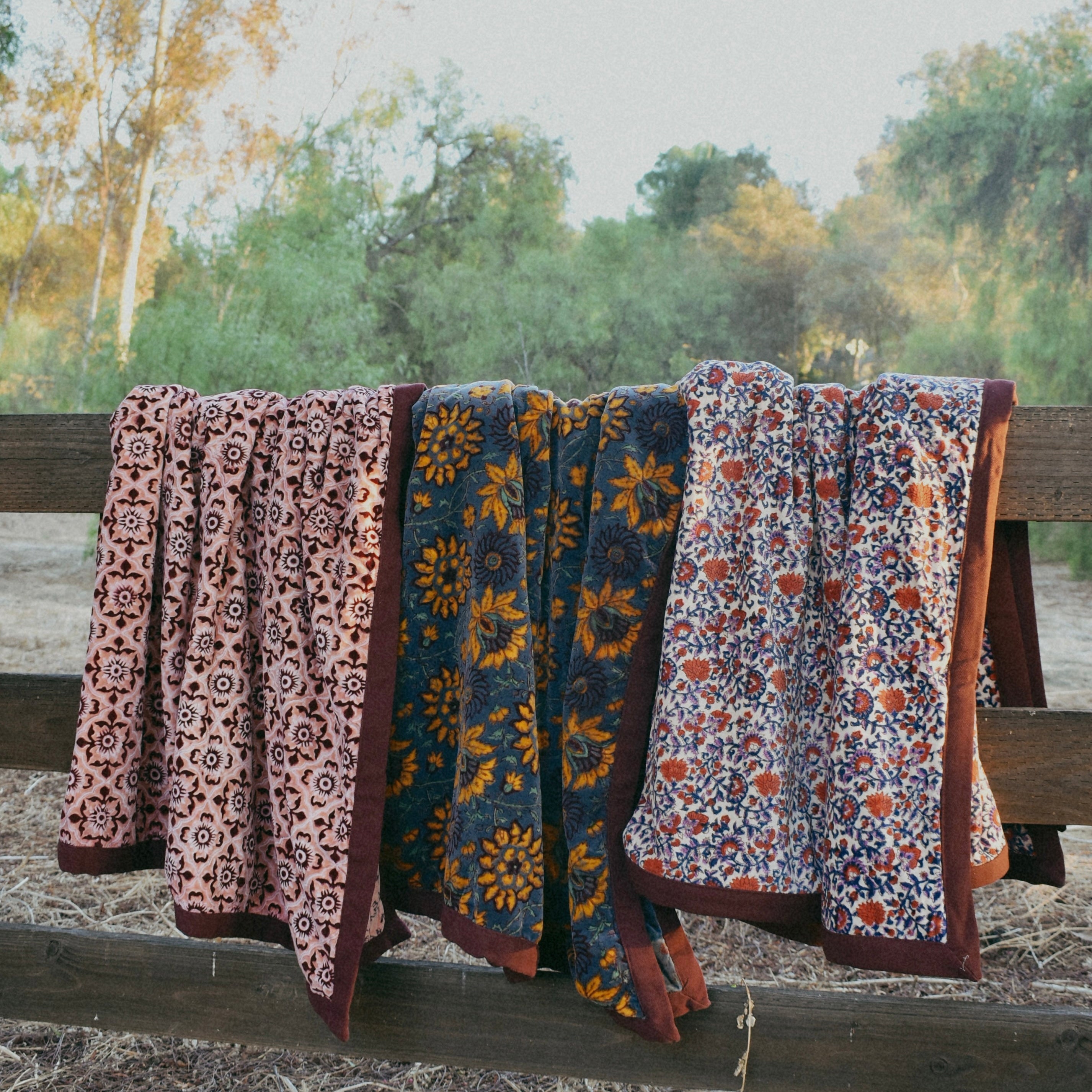 Three block printed velvet blankets, each with a unique all over floral print, draped over a wooden fence side by side next to each other with nature, trees in the background