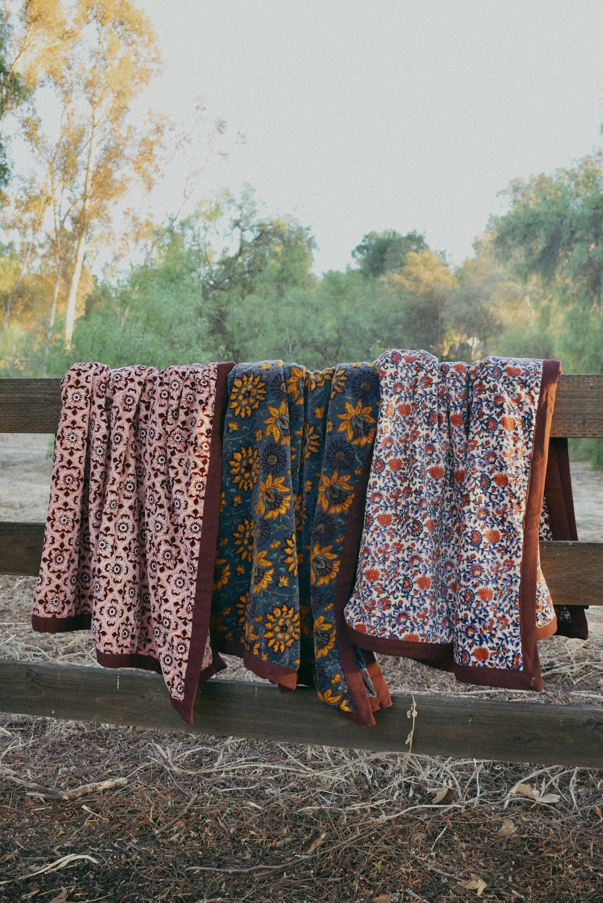 Three block printed velvet blankets, each with a unique all over floral print, draped over a wooden fence side by side next to each other with nature, trees in the background