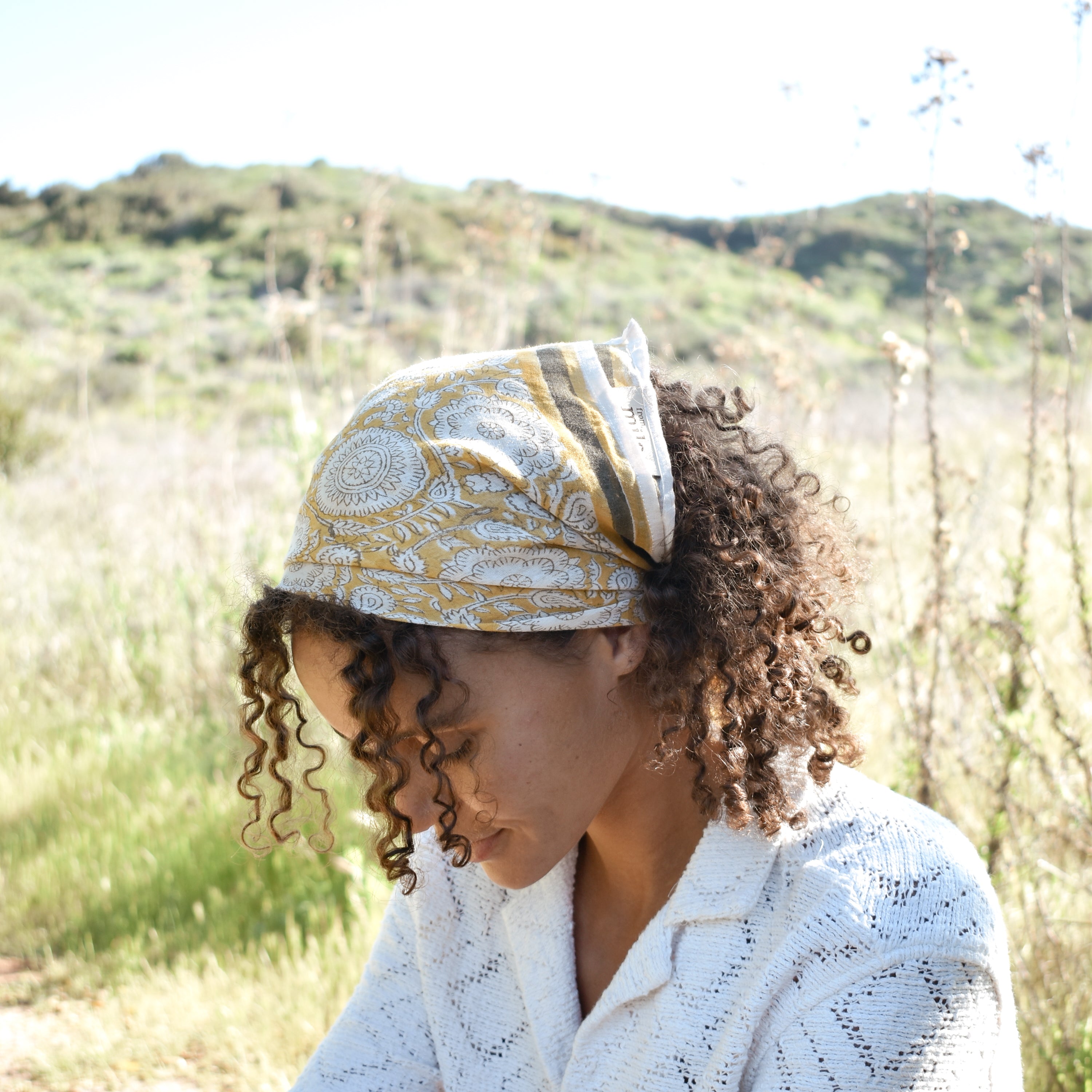 Woman wearing block print Tate bandana wrapped around the hair. Woman wearing a white, collared shirt. Foliage in the background.