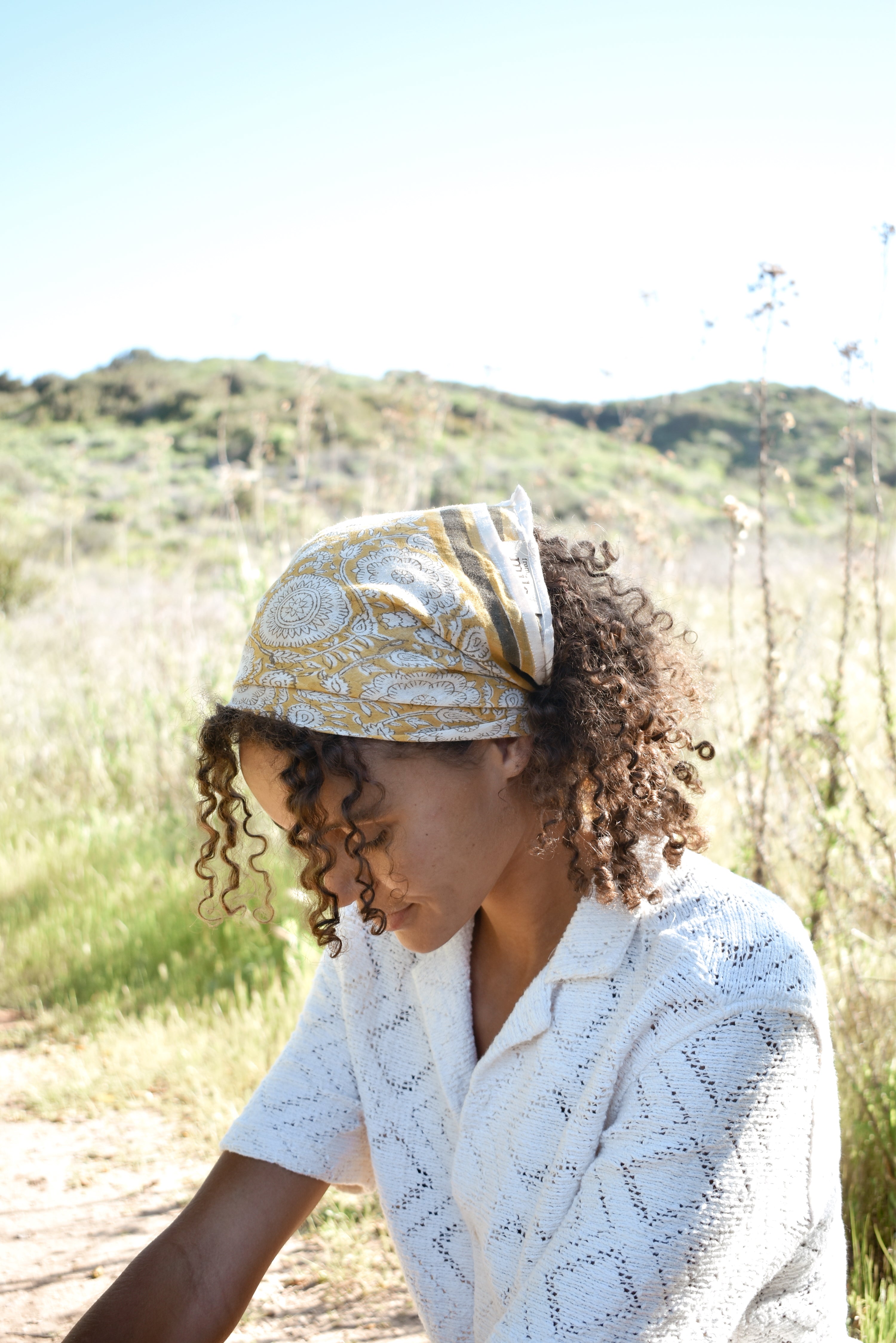 Woman wearing block print Tate bandana wrapped around the hair. Woman wearing a white, collared shirt. Foliage in the background.