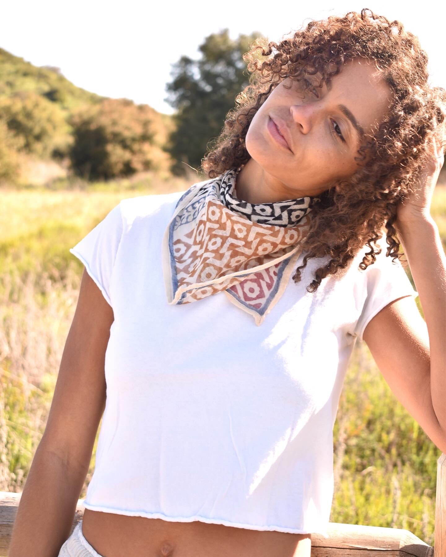 Woman in white tee wearing block printed Roma bandana around the neck, leaning against a wooden fence with trees in the background.
