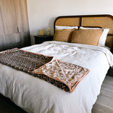 Block print velvet blanket. Tan background with brown diamond print. Reversible print has blue and gold diamond shapes. Displayed at the end of a bed. 