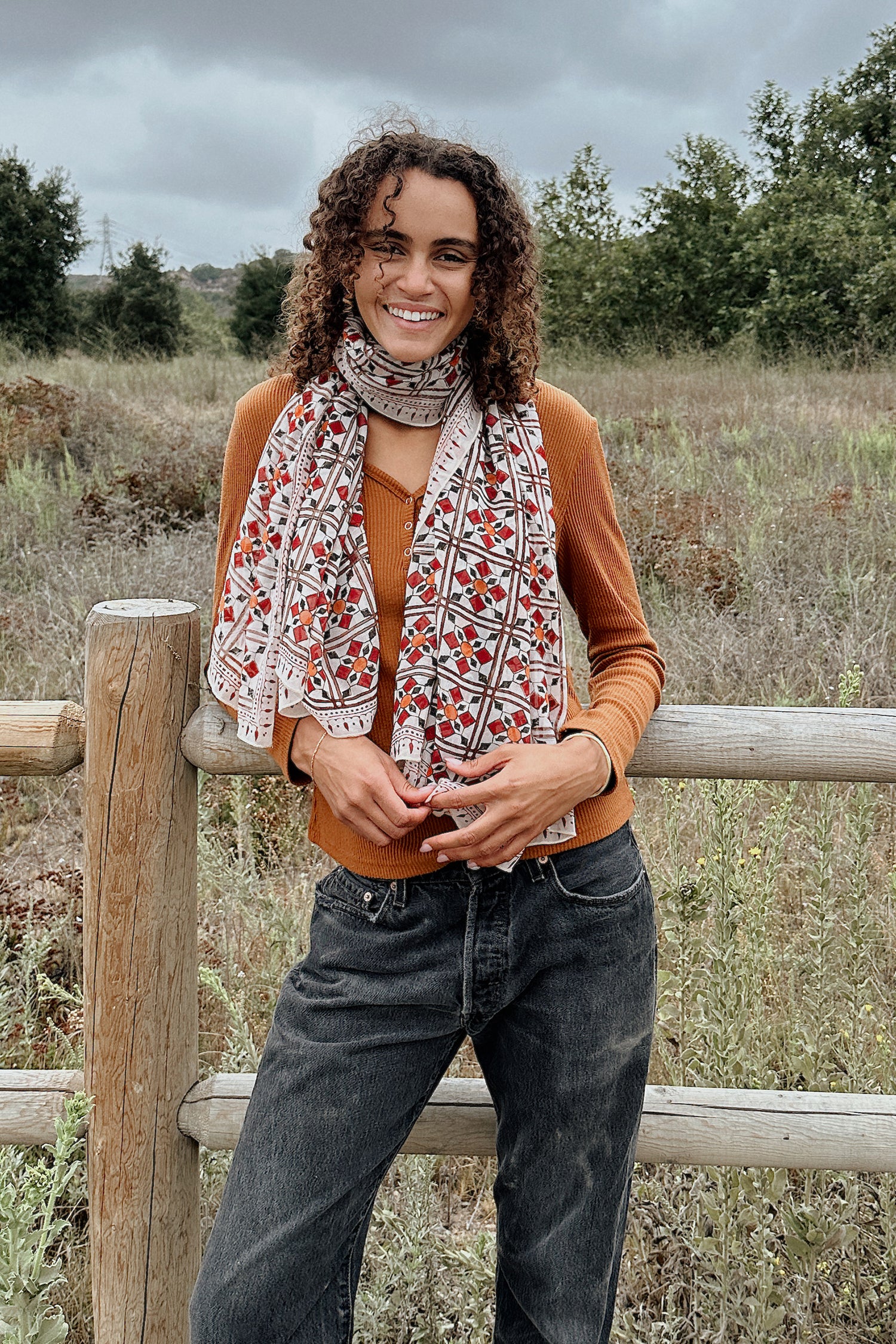 Woman wearing block printed Alia georgette scarf around the neck, paired with orange long sleeve and black jeans. Woman is standing outside in front of a fence.