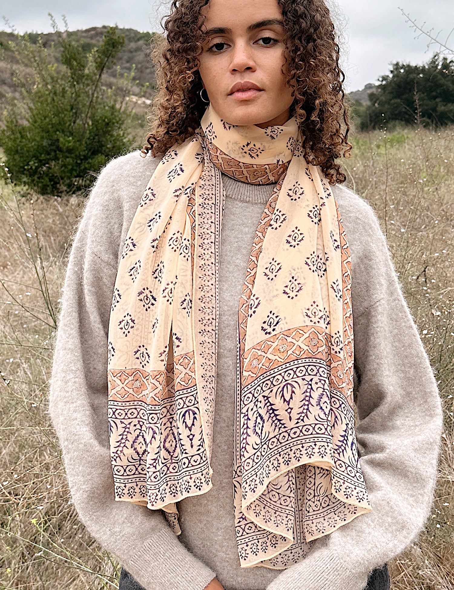Woman wearing block printed Alice georgette scarf around the neck, paired with tan sweater. Woman is standing outside.