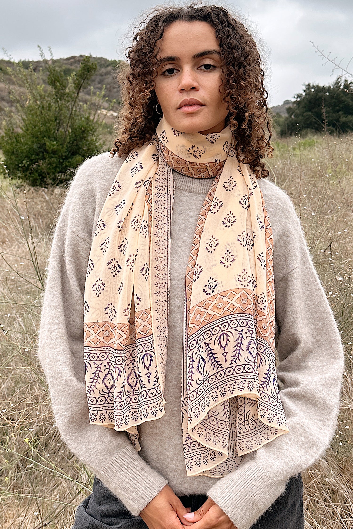 Woman wearing block printed Alice georgette scarf around the neck, paired with tan sweater. Woman is standing outside.