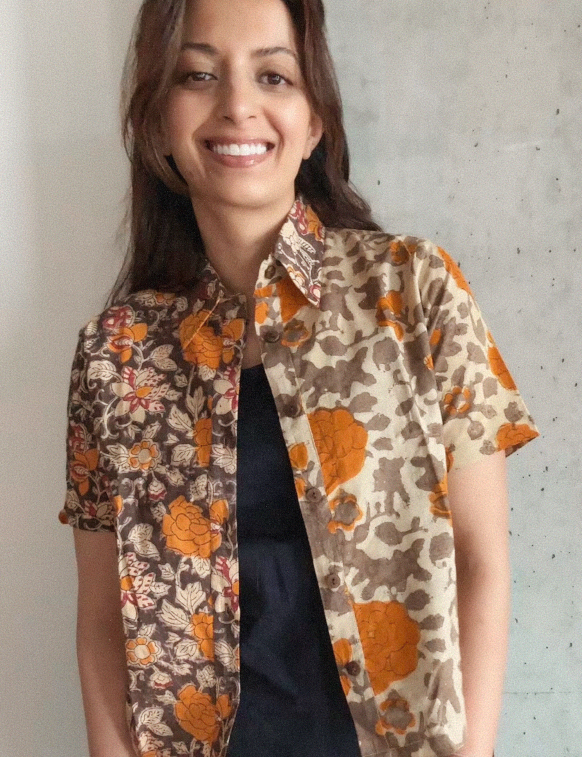 Woman wearing a grey and orange floral patterned shirt over a blakc dress against a concrete wall.