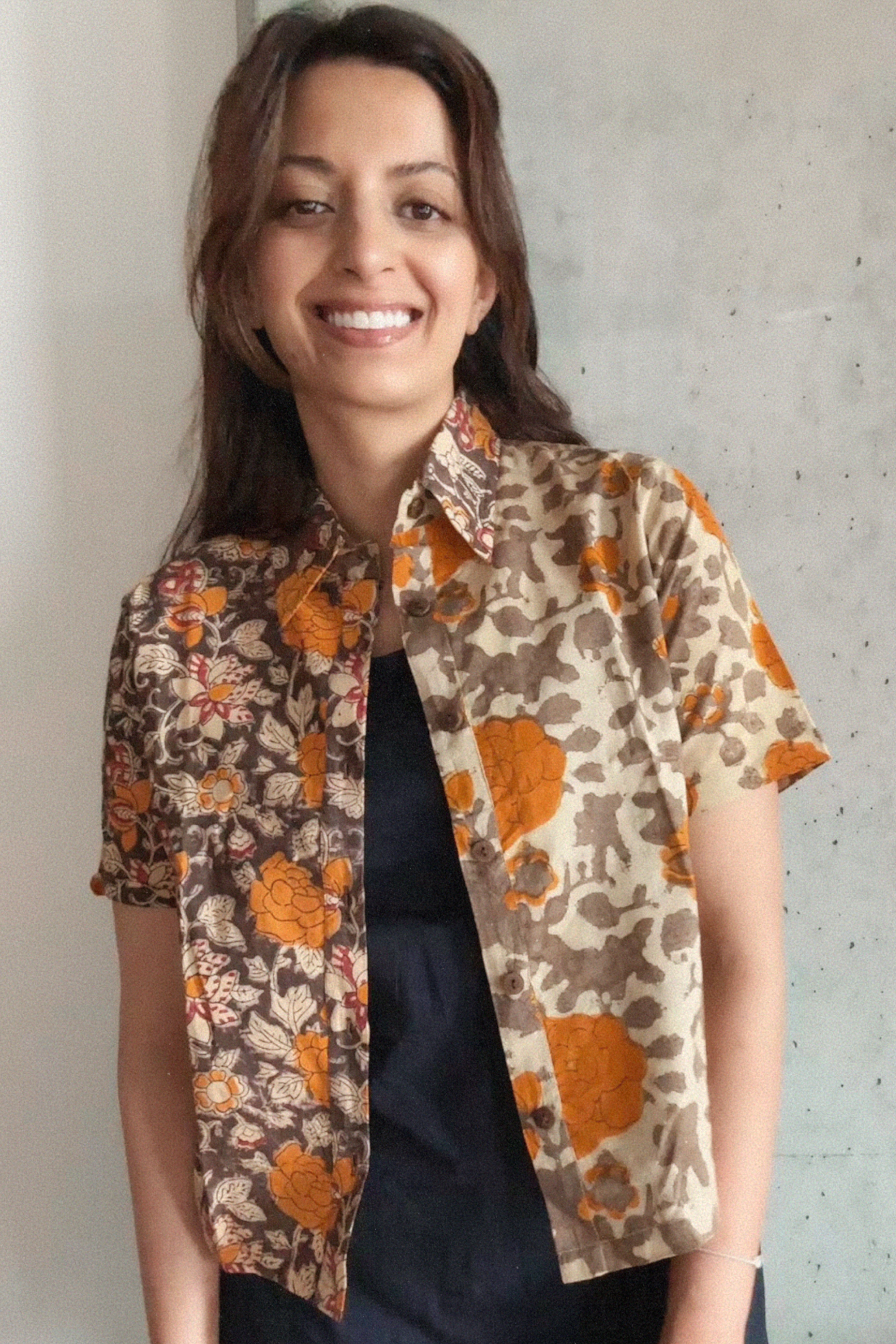 Woman wearing a grey and orange floral patterned shirt over a blakc dress against a concrete wall.