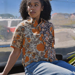 Woman in a floral shirt and jeans sitting outdoors in front of an RV