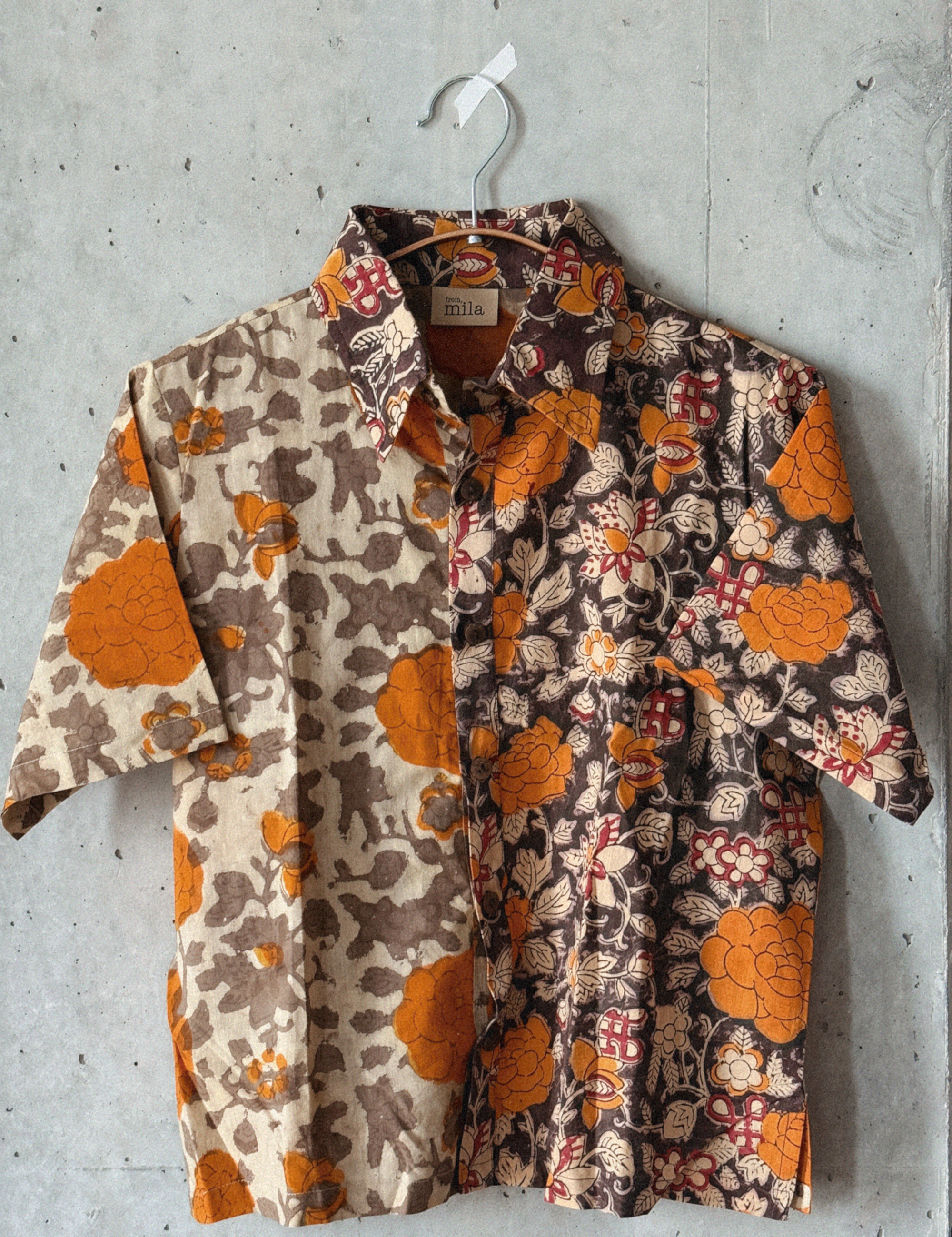 Orange and grey floral patterned shirt hanging on a hanger against a concrete wall.
