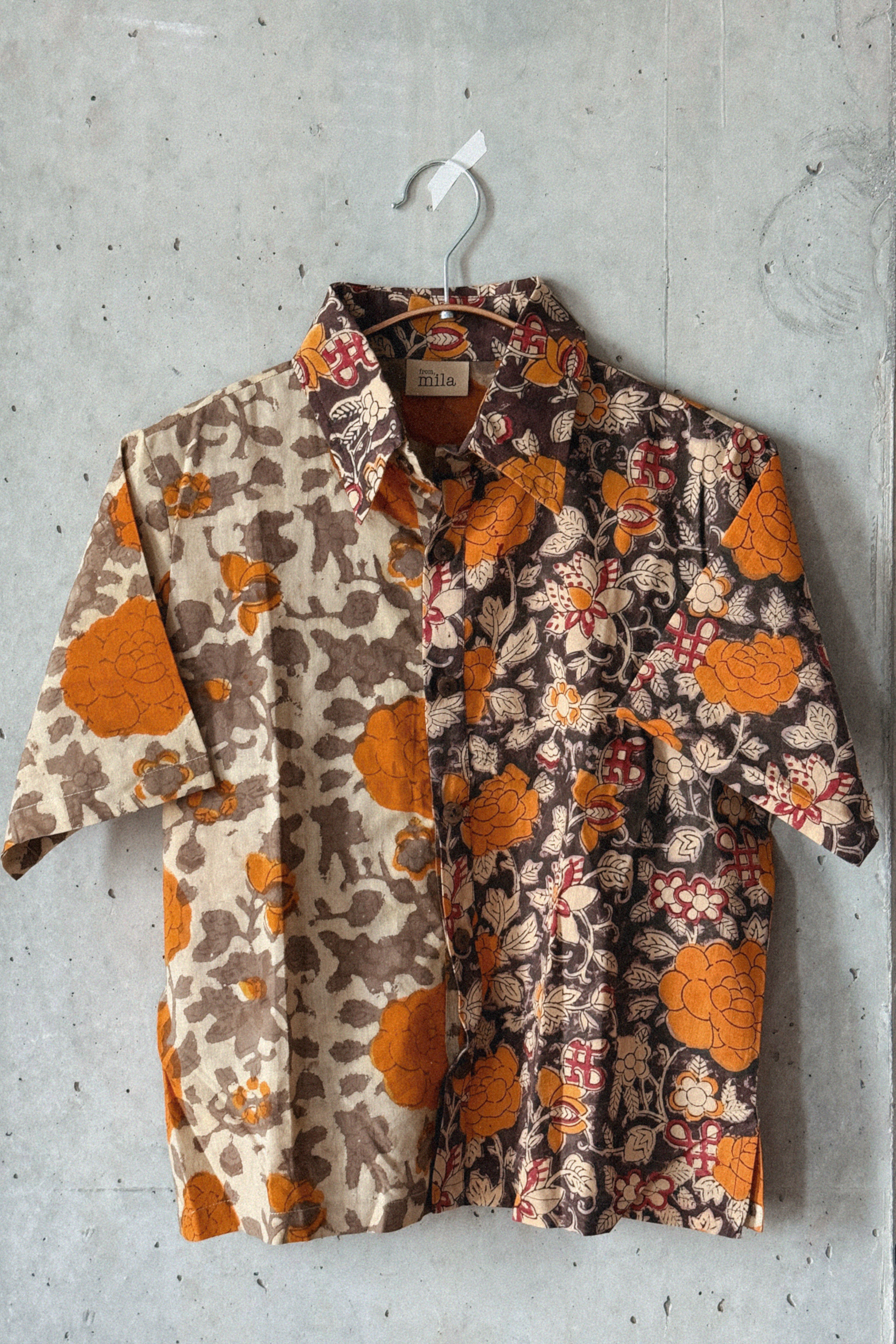 Orange and grey floral patterned shirt hanging on a hanger against a concrete wall.