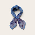 A knotted blue patterned scarf with paisley and polka dot motifs, tied in a loop against a light beige background.