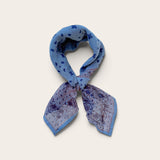 A knotted blue patterned scarf with paisley and polka dot motifs, tied in a loop against a light beige background.