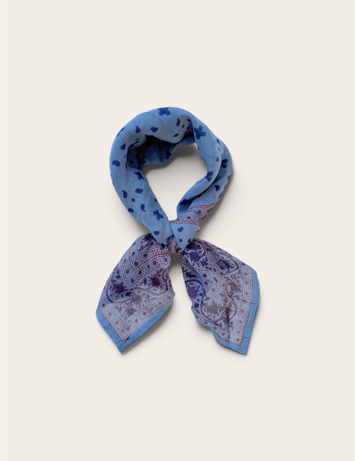 A knotted blue patterned scarf with paisley and polka dot motifs, tied in a loop against a light beige background.