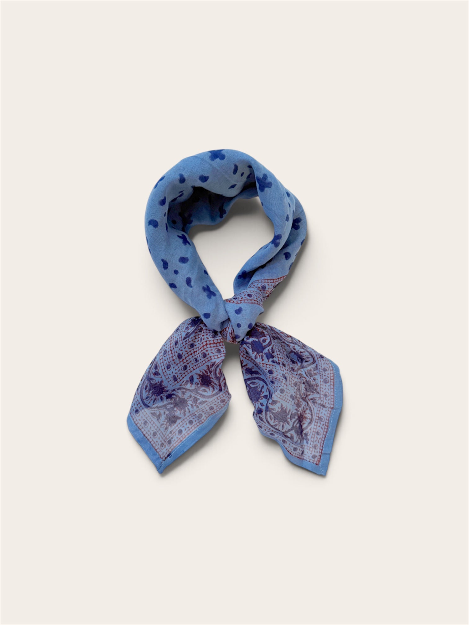 A knotted blue patterned scarf with paisley and polka dot motifs, tied in a loop against a light beige background.
